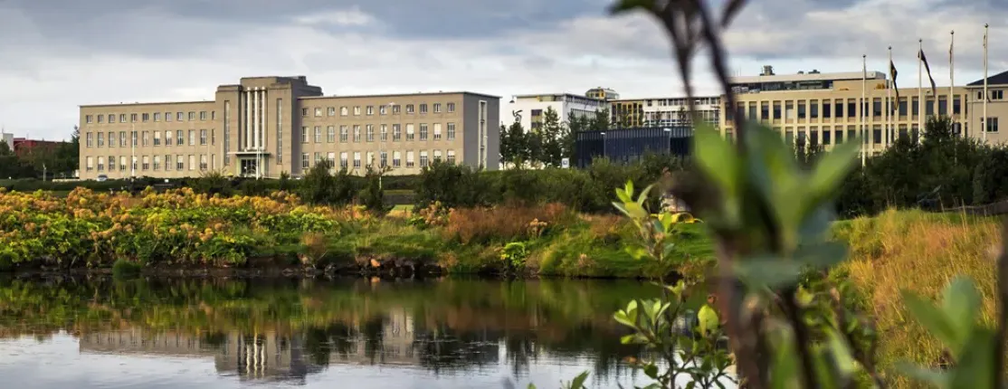 A scenic view of a large building with a pond in the foreground and greenery around, set against a cloudy sky